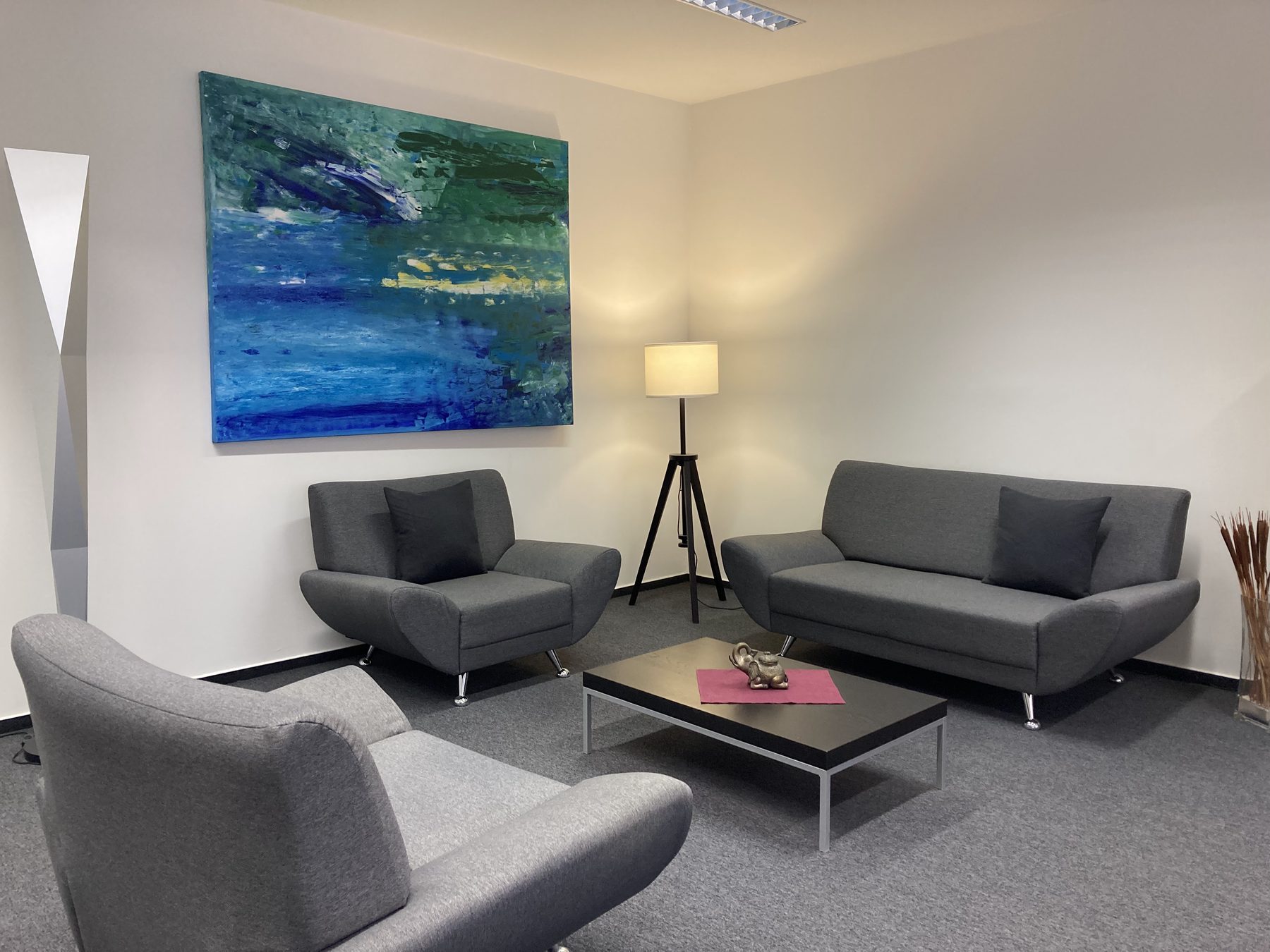 Der Behandlungsraum: zwei graue Sessel und ein Sofa um einen niedrigen Tisch, eine Stehlampe und ein großes blaugrünes Gemälde an der Wand.